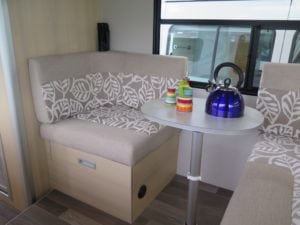 Motorhome seating in beige fabric with beige and white leaf patterned accent. Small round dining table with blue kettle and bright coloured tea set sit on table