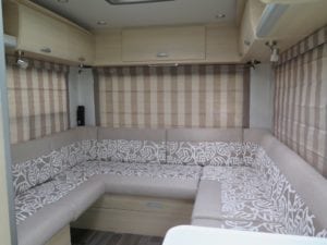 Motorhome interior with seating trimmed in two tone beige and white leaf pattern. Roman blinds on each side and at back are in beige and sand stripes in a closed position