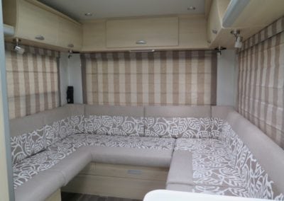 Motorhome interior with seating trimmed in two tone beige and white leaf pattern. Roman blinds on each side and at back are in beige and sand stripes in a closed position