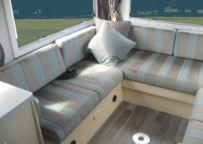 Motorhome seating in grey and blue stripe. Grey throw cushion sits in corner. Grass in background through window