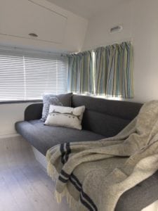 Large seat in caravan trimmed in charcoal with grey and white throw cushions and a draped throw. Closed venetian blinds and open striped curtains are in the background