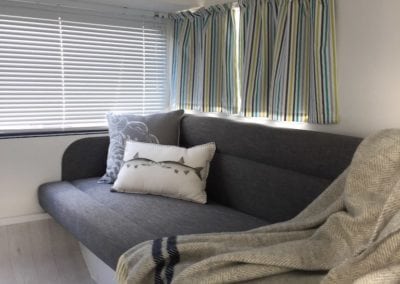 Large seat in caravan trimmed in charcoal with grey and white throw cushions and a draped throw. Closed venetian blinds and open striped curtains are in the background