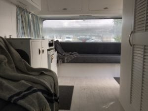 Caravan interior with a seat with a throw and cupboards in the foreground and large seat in front of a window in the background