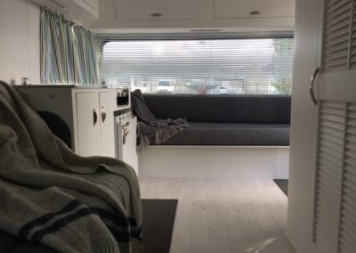 Caravan interior with a seat with a throw and cupboards in the foreground and large seat in front of a window in the background