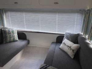 Two seats in caravan trimmed in charcoal. Small seat has a leaf patterned throw cushion. Large seat has grey and white throw cushions. Closed venetian blinds and open striped curtains are in the background