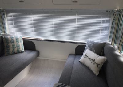 Two seats in caravan trimmed in charcoal. Small seat has a leaf patterned throw cushion. Large seat has grey and white throw cushions. Closed venetian blinds and open striped curtains are in the background