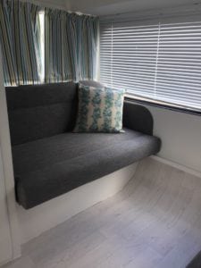 Small seat in caravan trimmed in charcoal with a leaf patterned throw cushion. Closed striped curtains sit behind and closed venetian blinds are to the right of the seat.