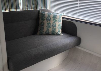 Small seat in caravan trimmed in charcoal with a leaf patterned throw cushion. Closed striped curtains sit behind and closed venetian blinds are to the right of the seat.