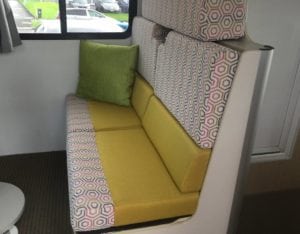Side view of motorhome dinette seating upholstered in yellow with a pink, grey, white geometric accent trim. A green throw cushion sits against the wall.