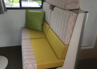 Side view of motorhome dinette seating upholstered in yellow with a pink, grey, white geometric accent trim. A green throw cushion sits against the wall.