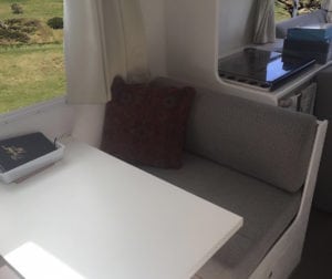 Dinette seating trimmed in beige with a wine coloured throw cushion. A white table with notebook in a small tray is in the foreground. Rural landscape can be seen through the window and more of the motorhome in the background.