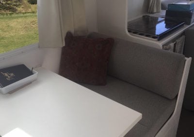 Dinette seating trimmed in beige with a wine coloured throw cushion. A white table with notebook in a small tray is in the foreground. Rural landscape can be seen through the window and more of the motorhome in the background.