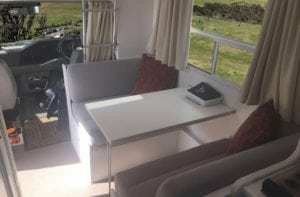 Refurbished dinette seating trimmed in beige. Wine coloured throw cushions accessorise each seat. A white table with a notebook in small tray sits on the table. A rural setting can be seen through the side window. The drivers cabin can be seen in the background.