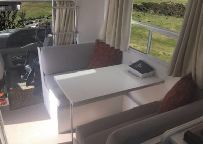 Refurbished dinette seating trimmed in beige. Wine coloured throw cushions accessorise each seat. A white table with a notebook in small tray sits on the table. A rural setting can be seen through the side window. The drivers cabin can be seen in the background.