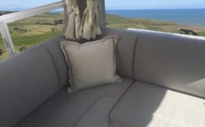 Corner of motorhome seating in beige trim with cream throw cushion in corner. Rural setting and seascape can be seen in background