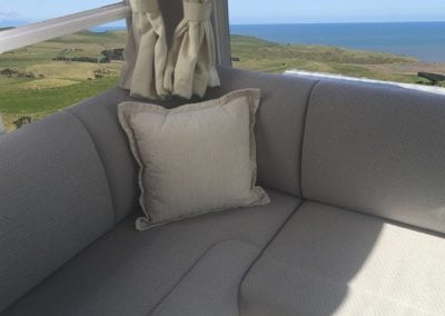 Corner of motorhome seating in beige trim with cream throw cushion in corner. Rural setting and seascape can be seen in background