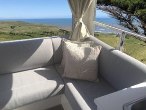 Motorhome interior with seating trimmed in beige. Cream curtains and cream throw cushions accessorise the lounge cushions. Rural background and seascape are in background through the corner window.