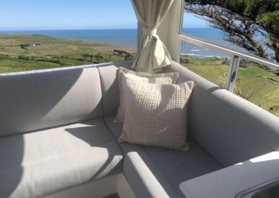 Motorhome interior with seating trimmed in beige. Cream curtains and cream throw cushions accessorise the lounge cushions. Rural background and seascape are in background through the corner window.