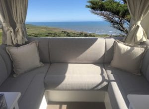 Motorhome seating in beige trim. Cream throw cushions sit in each corner. Rural hills and seascape is in background through rear window