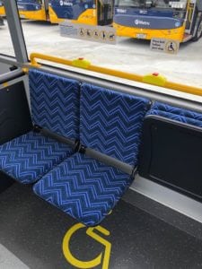 Mobility bus seating installed in bus trimmed in blue chevron pattern. Left hand and middle seats in open position. Right hand seat in closed position. Buses can be seen in background through side window.