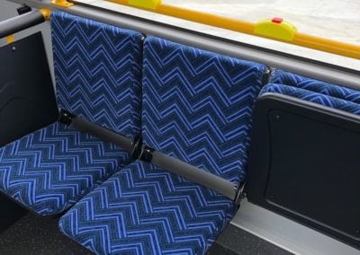 Mobility bus seating installed in bus trimmed in blue chevron pattern. Left hand and middle seats in open position. Right hand seat in closed position. Buses can be seen in background through side window.