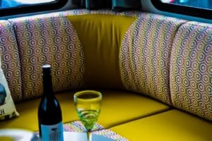 Motorhome corner lounge cushions in pink, white and grey geometric pattern and plain bright yellow. Bottle of wine and wine glass are in the foreground.