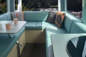 Motorhome lounge with cushions in a soft green shade. Throw cushions sit in each corner of the lounge. Table with dinnerware sits to the left hand side. Scenery can be seen through the windows.