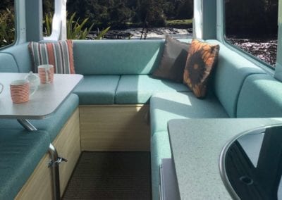 Motorhome lounge with cushions in a soft green shade. Throw cushions sit in each corner of the lounge. Table with dinnerware sits to the left hand side. Scenery can be seen through the windows.