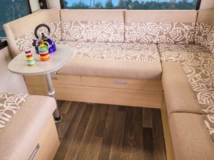Motorhome lounge two tone cushions with beige and white leaf pattern and beige plain fabric on edges. Table with kettle and dinnerware sit to the left hand side of the lounge