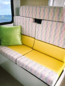 Motorhome dinette seating upholstered in yellow with a pink, grey, white geometric accent trim. A green throw cushion sits against the wall.