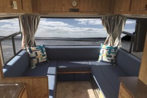 Motorhome lounge with dark blue wrap around seat and back cushions. Seat cushion has pull down detailing. Beach can be seen in background through rear window.