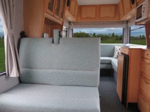 Front facing dinette in grey green fabric. Single cushion with no head rest. Pull down detail on both seat and back cushion. Lounge is in background. Grass can be seen through rear window.